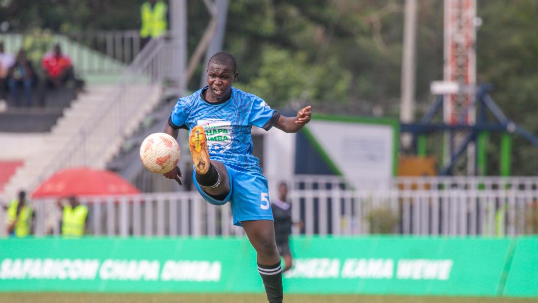 A footballer in a blue number five kit raises a boot towards the ball on a pitch with Safaricom Chapa Dimba branding.