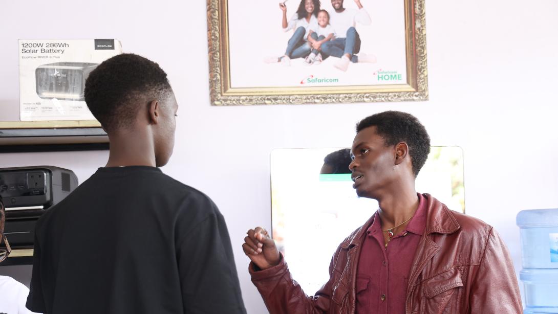 Two men converse beside equipment displays and Safaricom Home branding, with one gesturing while holding a phone and leaflet.
