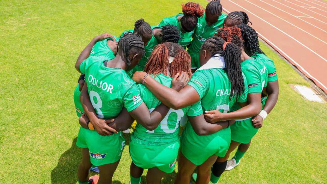 Women rugby players in green jerseys huddle on a grass pitch beside a stadium running track.