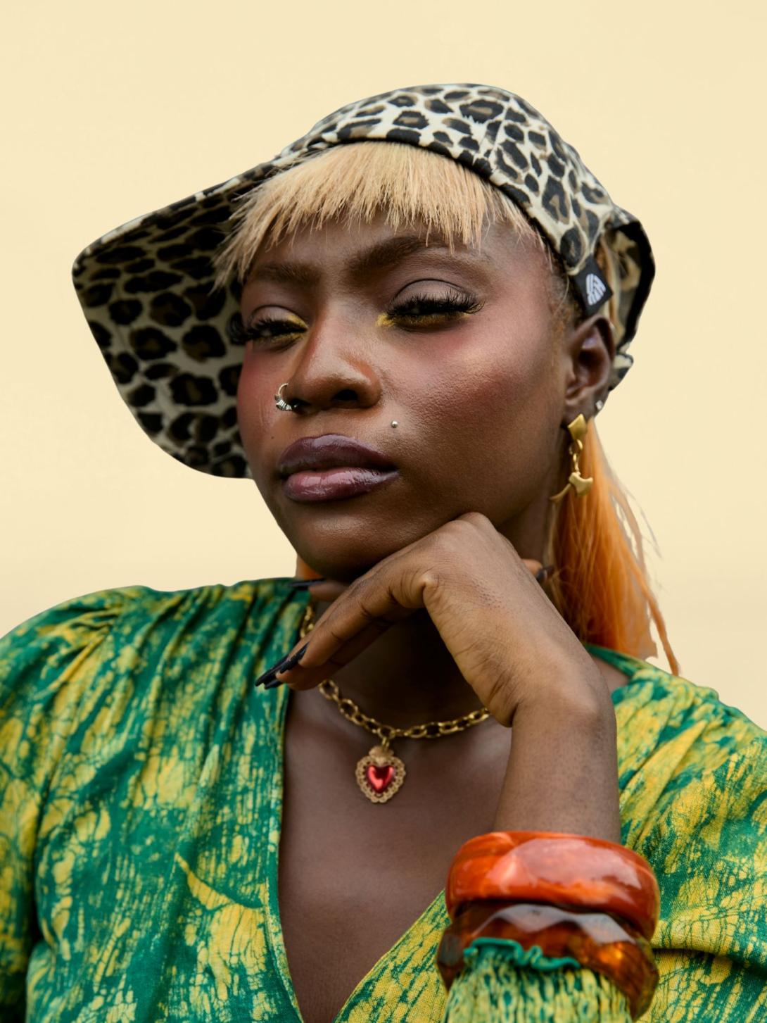 Nigerian singer and songwriter Solana poses in a leopard-print cap and green patterned outfit, resting her chin on her hand.