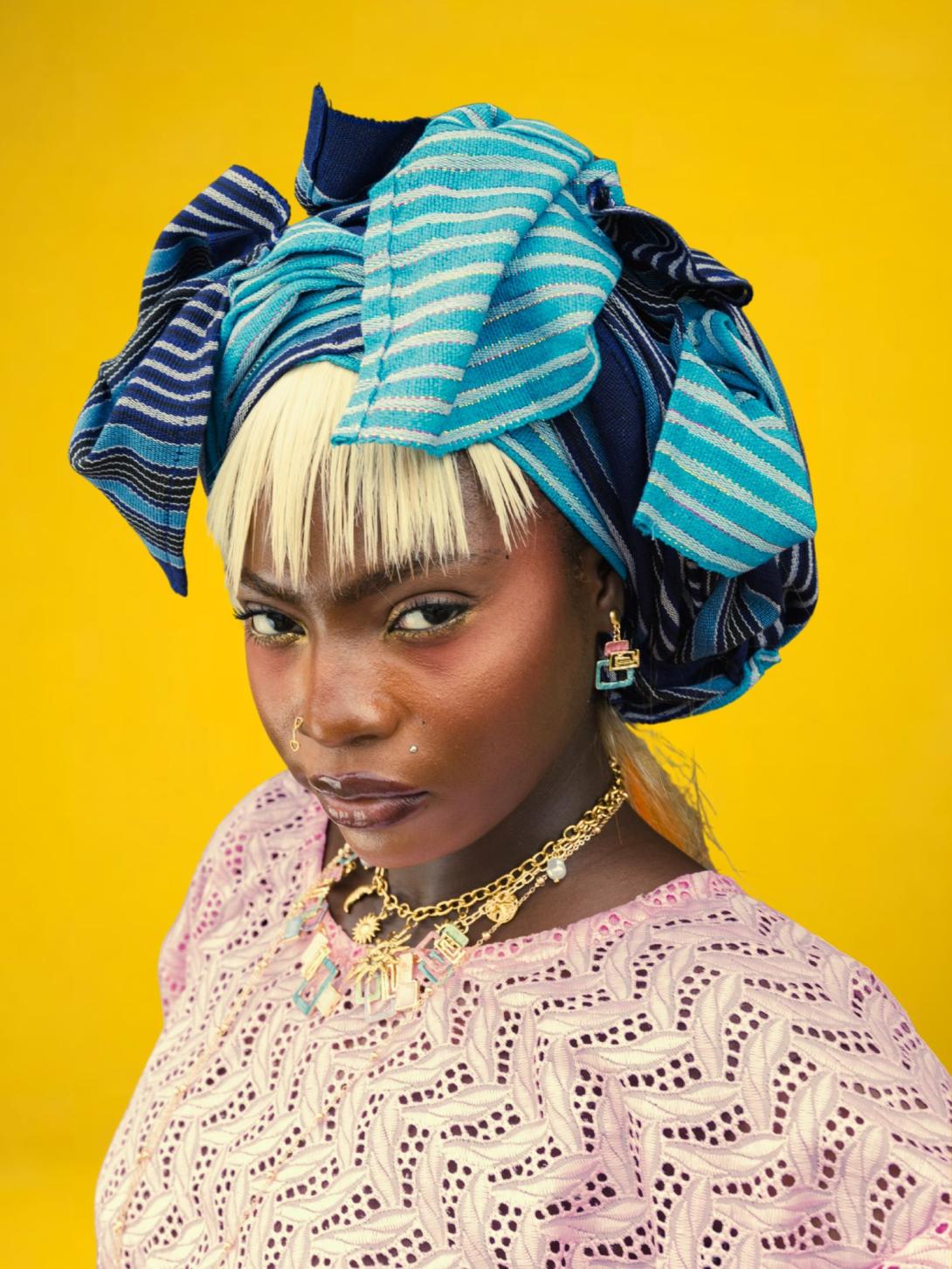 Nigerian singer and songwriter Solana wears a blue striped headwrap, pink lace outfit and layered necklaces against a yellow background.