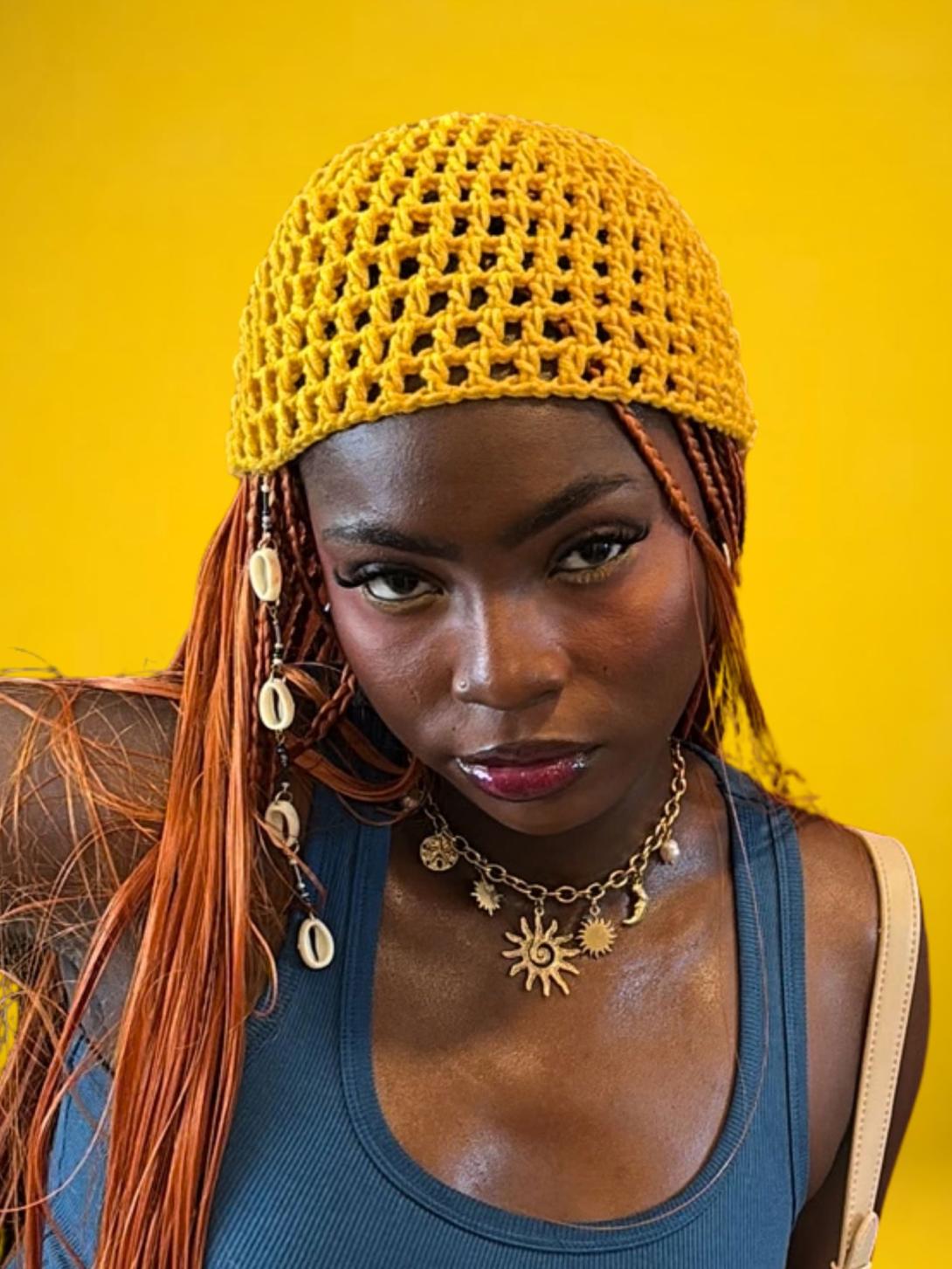 A woman wearing a yellow crocheted cap, shell-adorned braids and a sun pendant necklace poses against a yellow background.