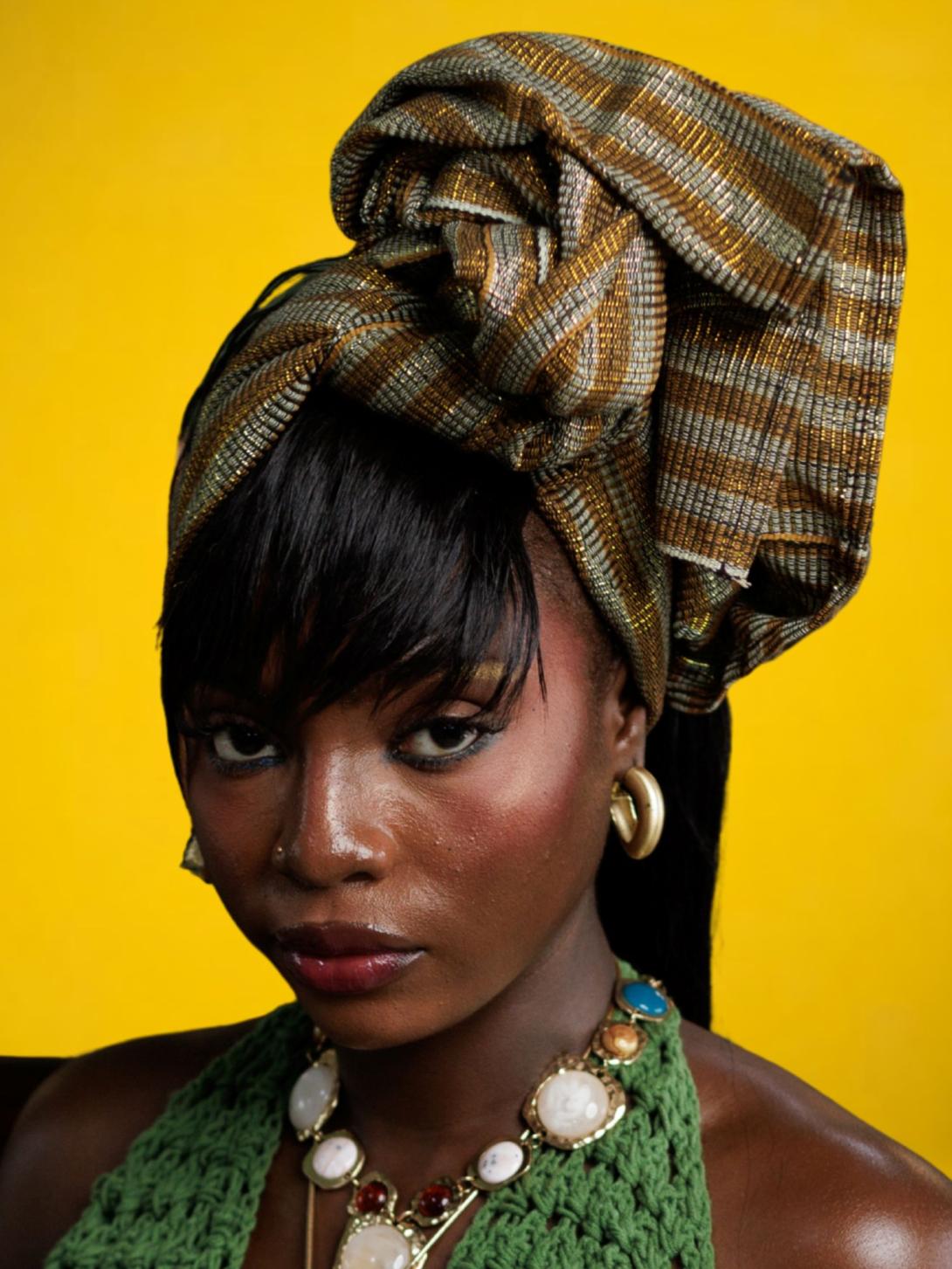 A woman wearing a patterned headwrap, green crocheted top and statement jewellery poses against a yellow background.