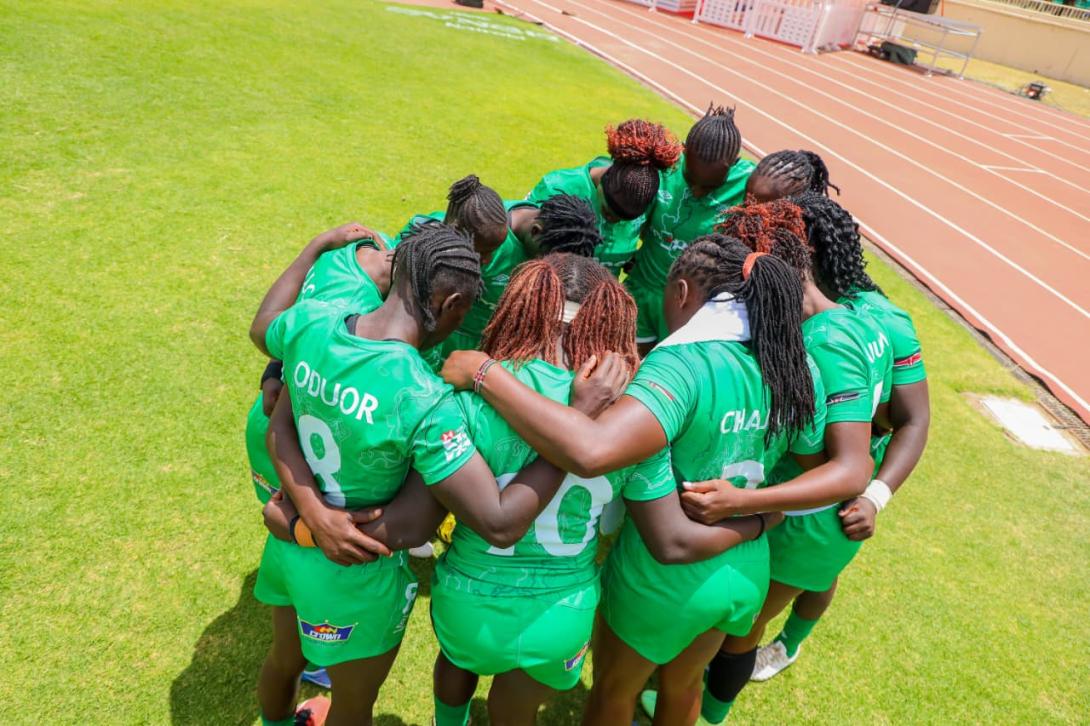 Women rugby players in green Kenya kits form a huddle on a grass pitch beside a running track.