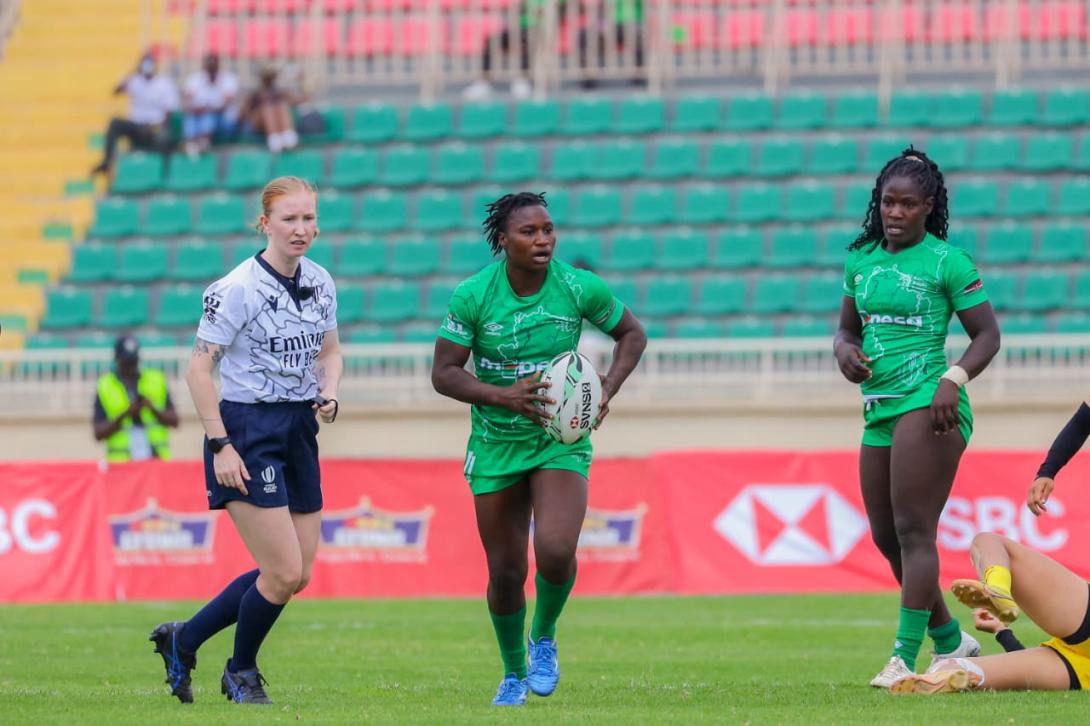 A rugby player in a green M-PESA-branded kit carries the ball during a match, with a teammate and referee nearby.