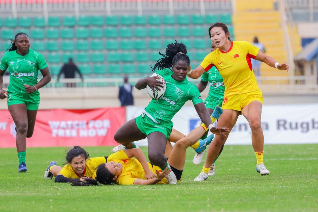A rugby player in a green M-PESA-branded kit carries the ball as opponents in yellow attempt to tackle her.