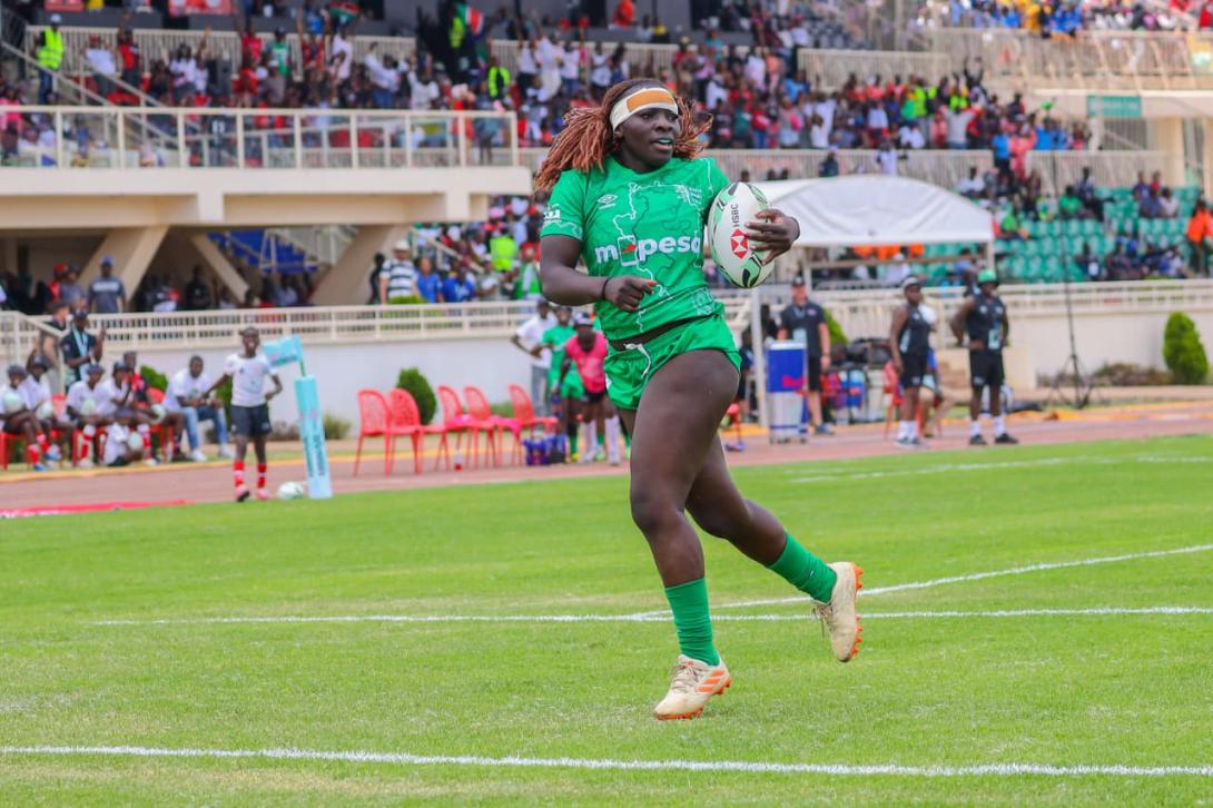 A rugby player in a green M-PESA-branded kit runs with the ball as spectators watch from the stadium stands.