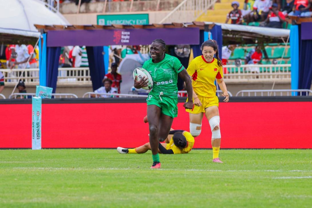 A rugby player in a green M-PESA-branded kit carries the ball as an opponent in yellow chases her.