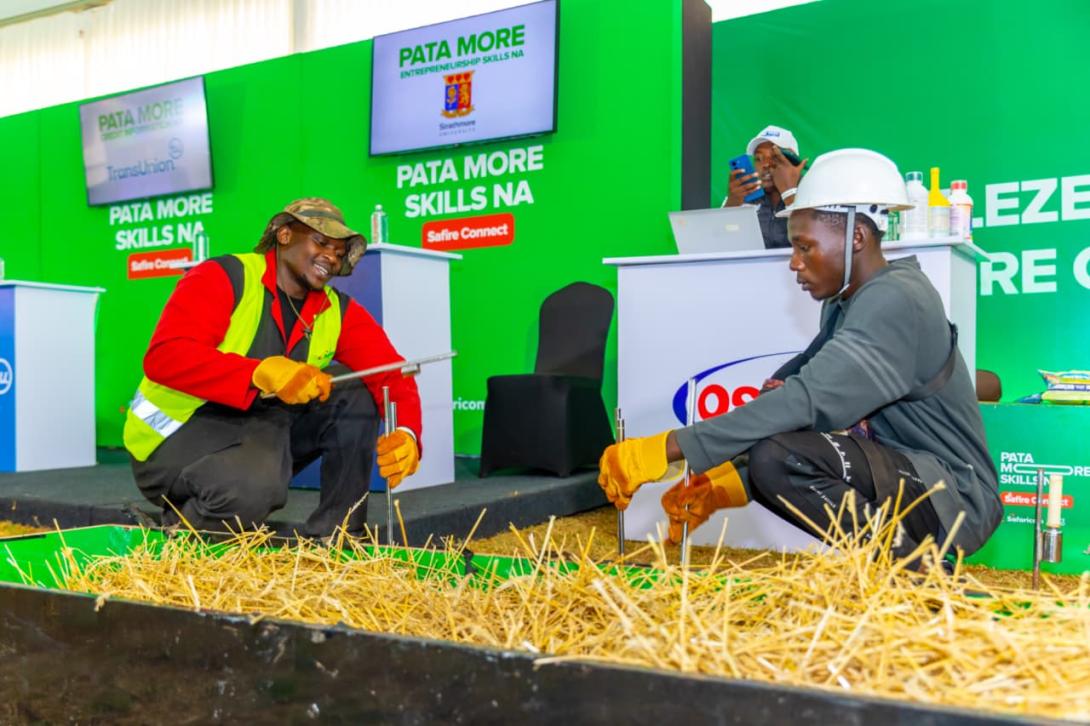 Two men wearing protective gloves handle metal tools beside a straw-filled display at Safaricom’s Safire Connect forum in Githunguri.